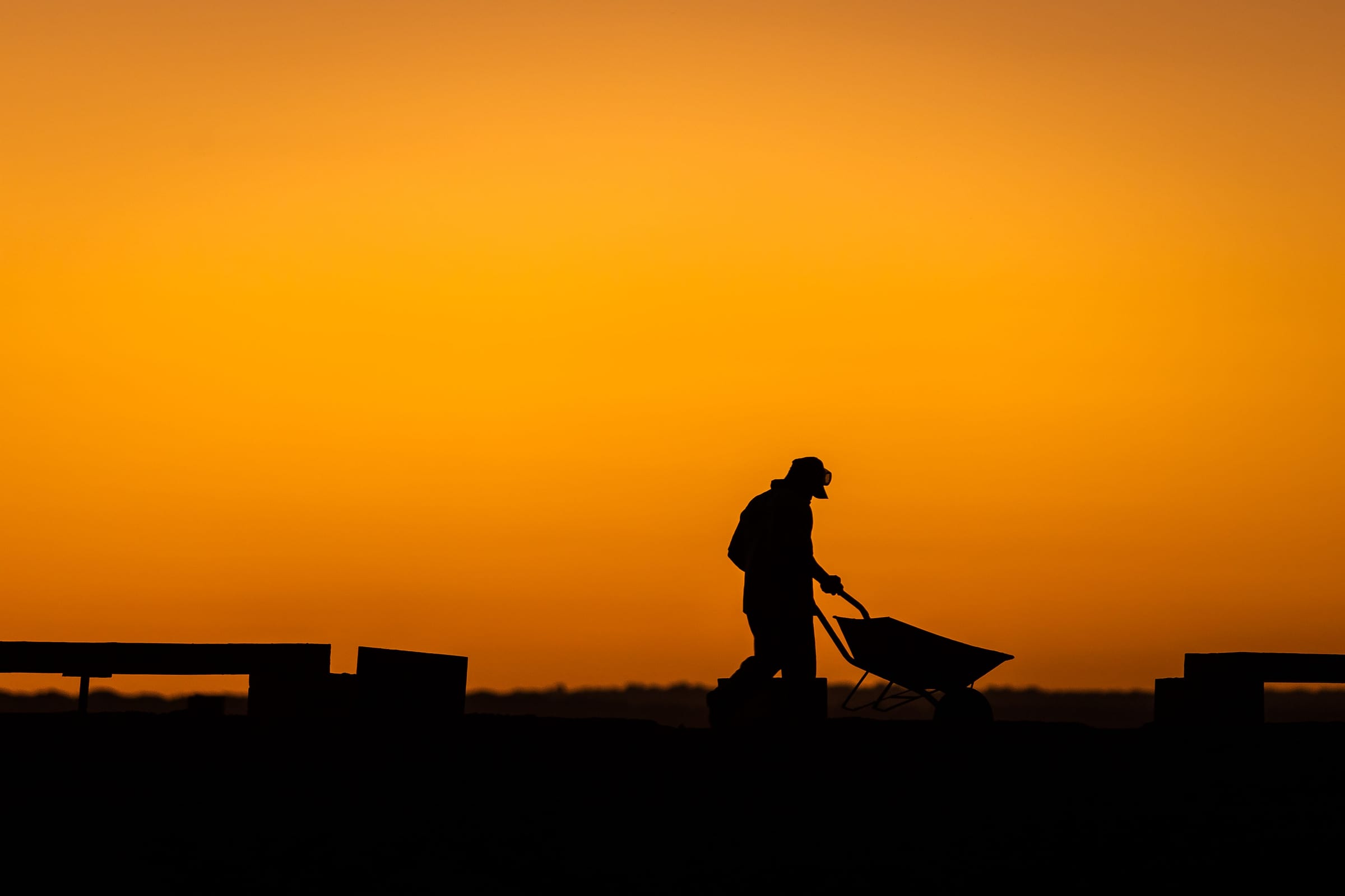 A construction worker silhouetted against a sunset, pushing a wheelbarrow.